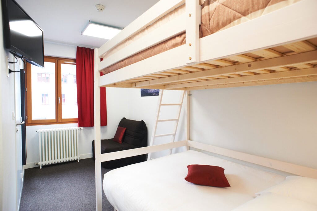 Une chambre d'hôtel lumineuse et propre avec un lit superposé. Le lit du bas a une literie blanche et un oreiller rouge, et le lit du haut également. Une fenêtre laisse entrer la lumière naturelle.